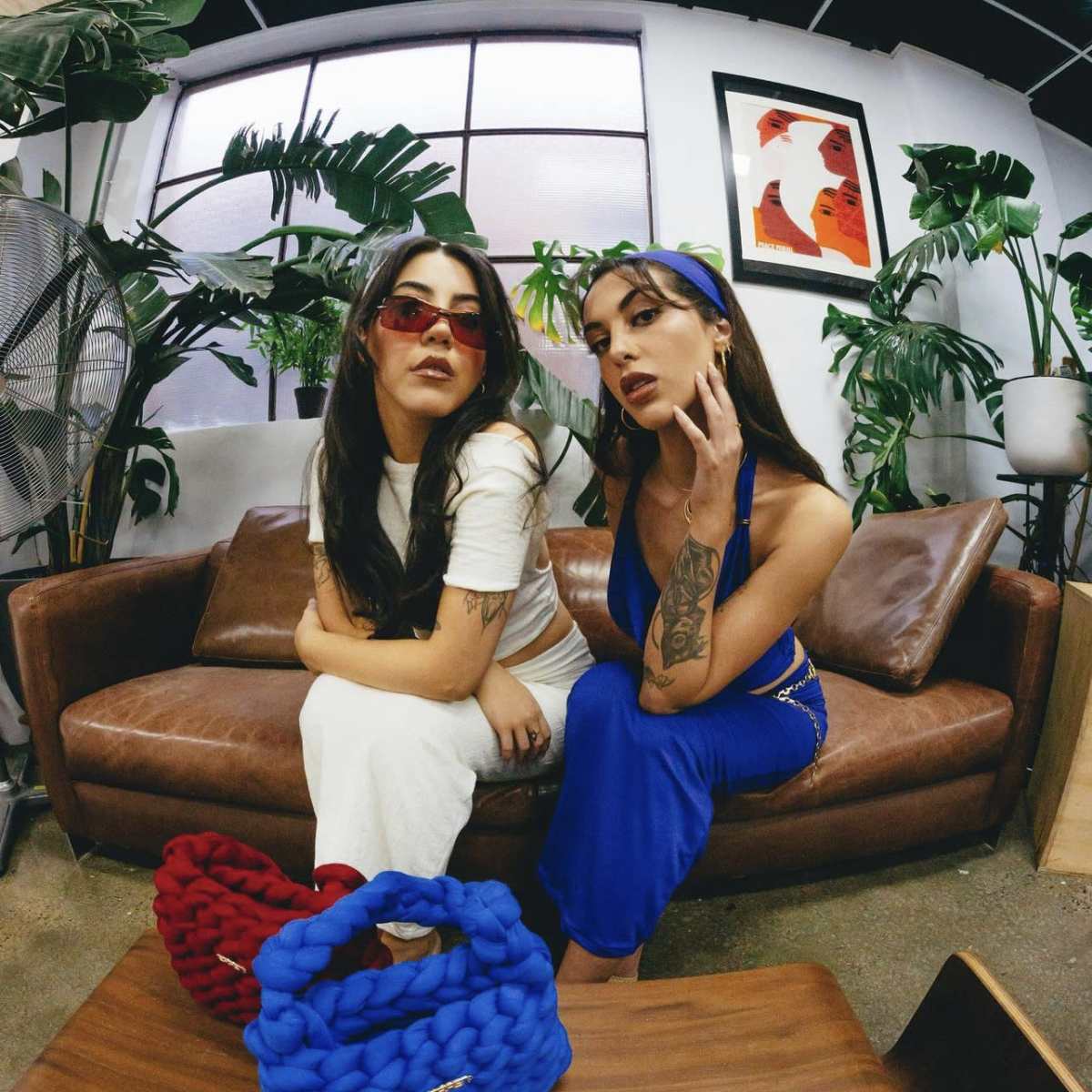 Two women pose on a brown leather sofa in a green, plant-filled room, wearing colorful outfits and accessories.