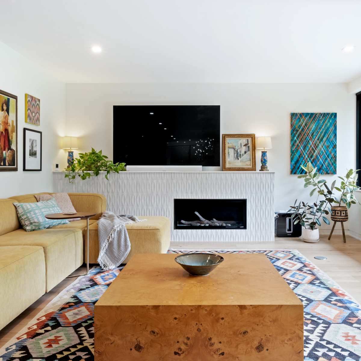 A modern living room features a yellow sectional sofa, a rectangular wooden coffee table, framed artwork on the walls, and a wall-mounted television above a fireplace.