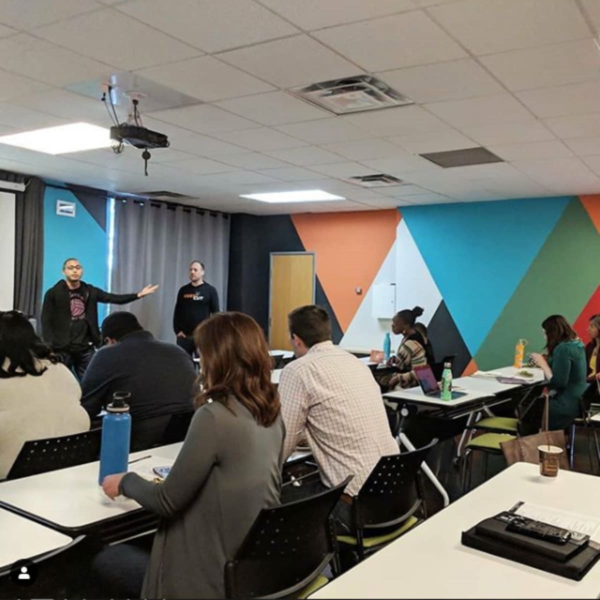 Two speakers are presenting in front of a seated audience in a brightly colored classroom.