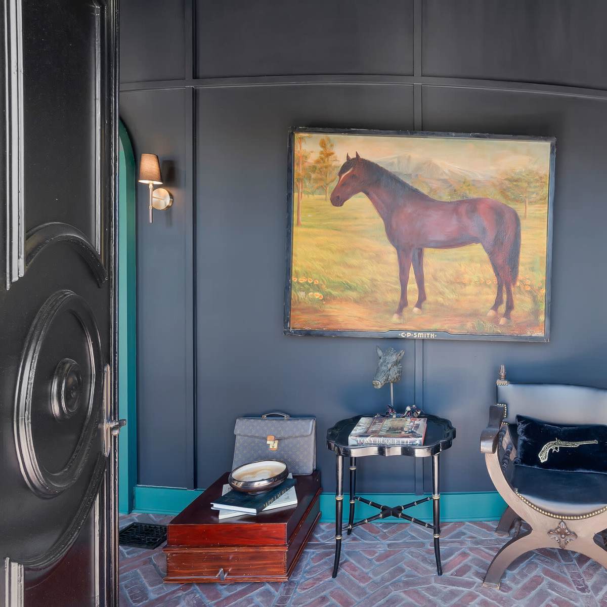 A small room with a painting of a horse on the wall, a chair, a side table, and a suitcase on the floor.