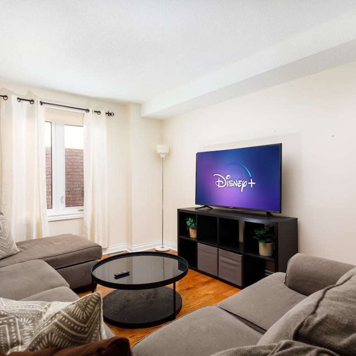 A living room features a gray sectional sofa, a round coffee table, and a television displaying the Disney+ logo, with a window covered by sheer curtains.