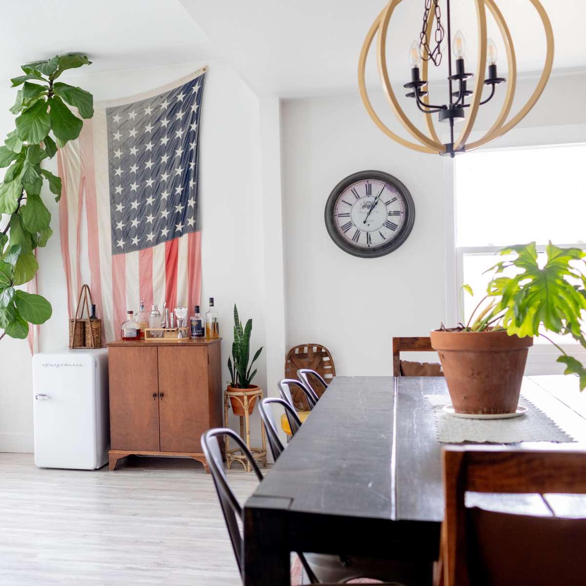 A spacious dining area features a black wooden table, a vintage clock on the wall, a potted plant beside a flag decor, and a small refrigerator near a wooden cabinet.