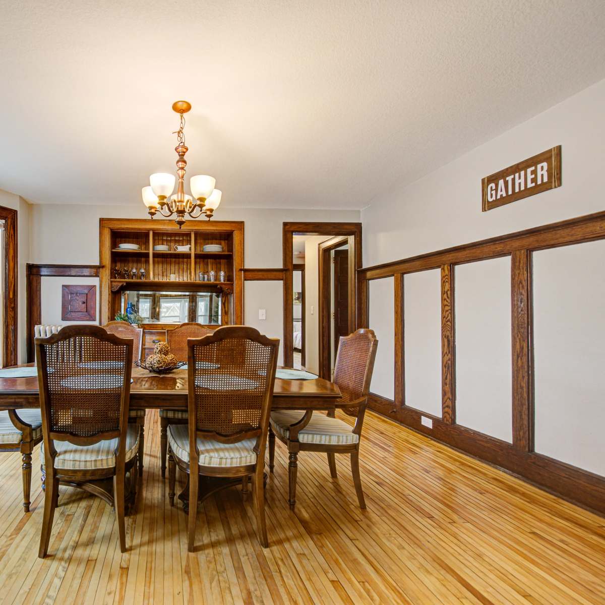 A dining room features a wooden table surrounded by chairs, with large windows allowing natural light and a chandelier hanging above.
