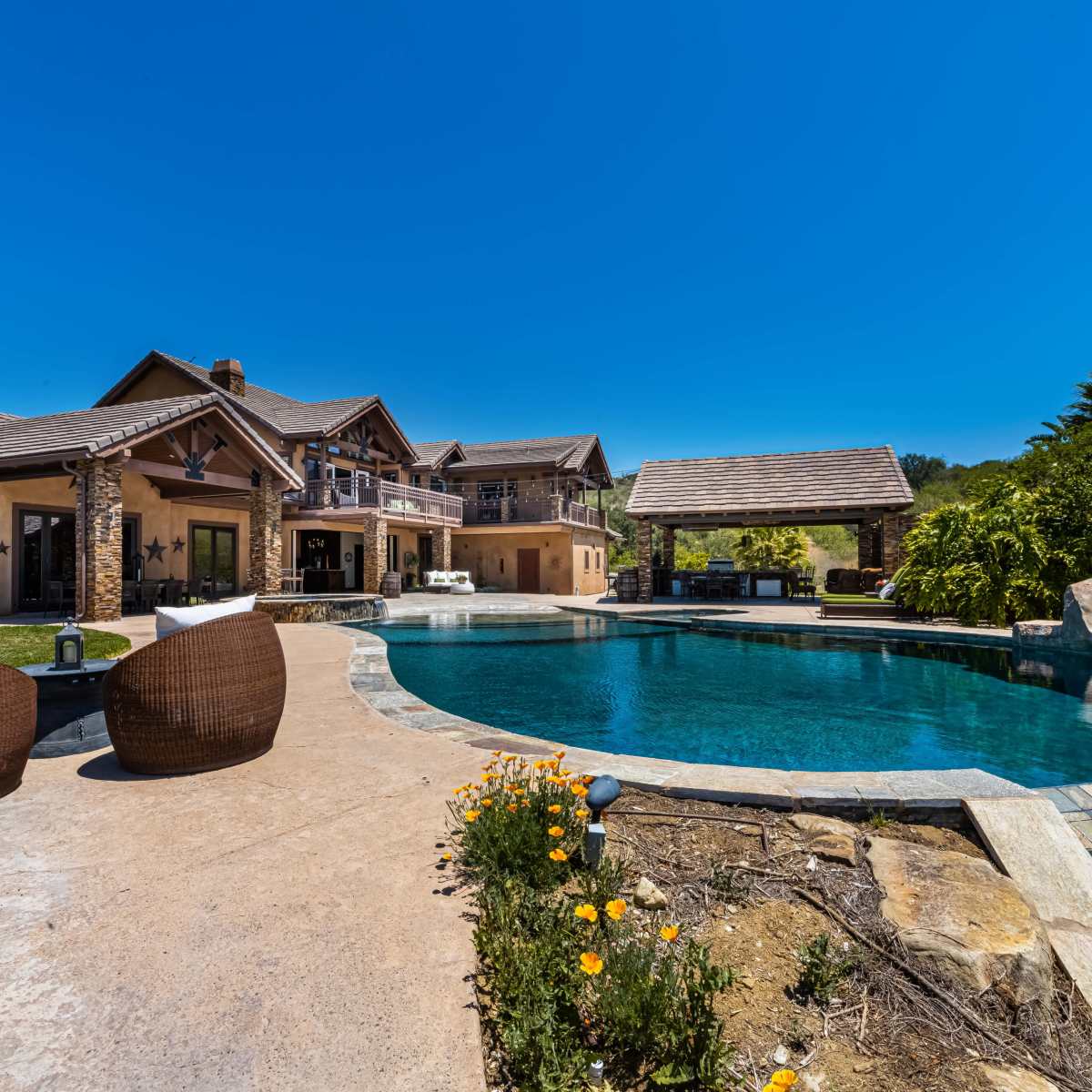 A spacious backyard featuring a swimming pool with a slide, surrounded by a modern house and lush greenery under a clear blue sky.