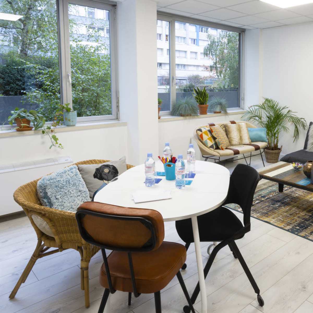 A bright office lounge area with a black sofa, a round white dining table with chairs, and several potted plants.