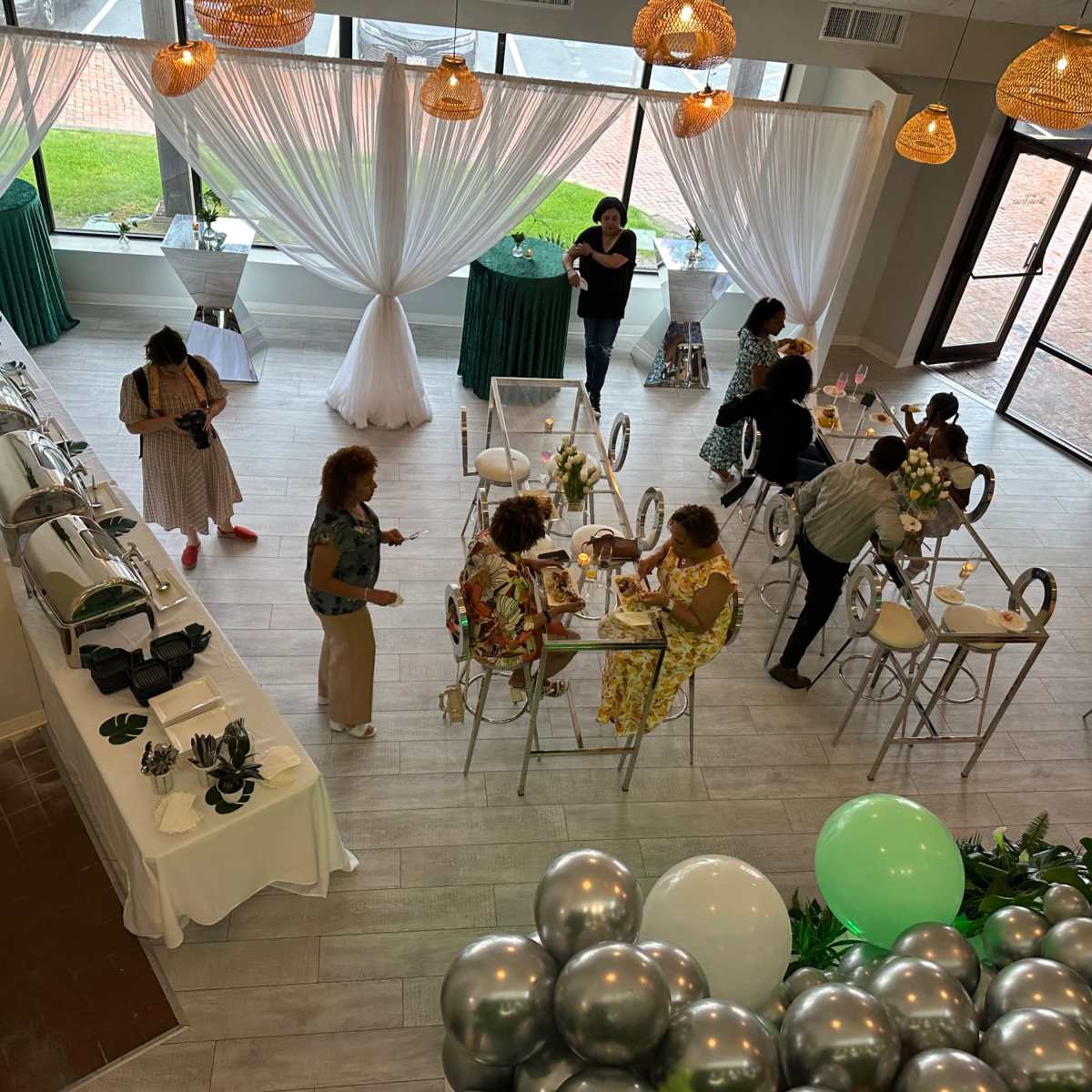 A bright event space with tables set for dining, guests gathering, and decorative elements like balloons and hanging lights.