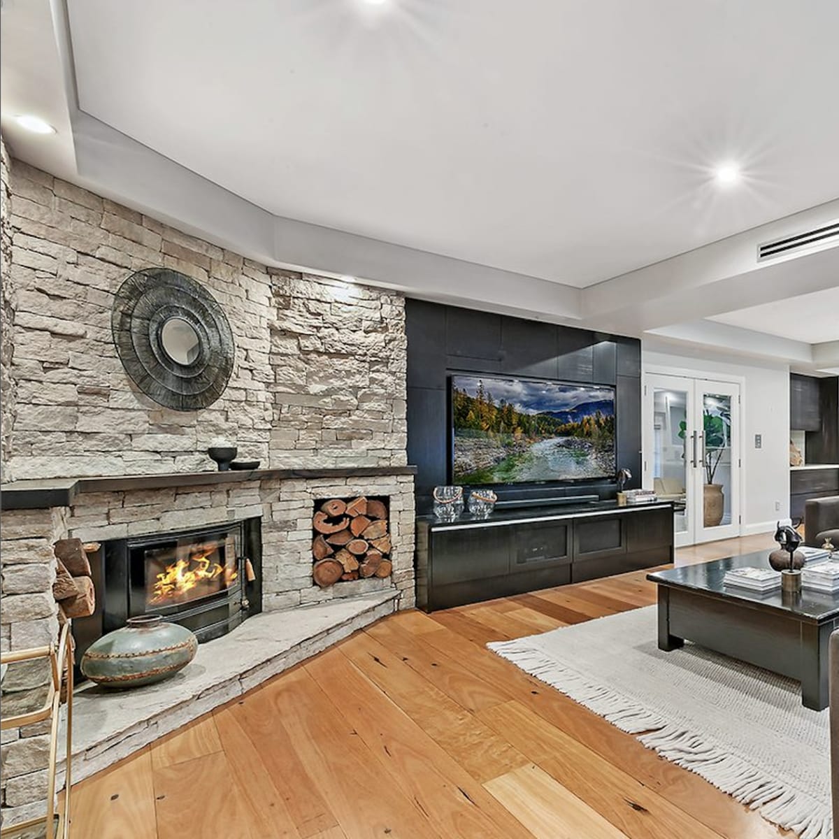 A modern living room featuring a stone fireplace, a large television on a dark cabinet, and a sectional sofa arranged around a coffee table.