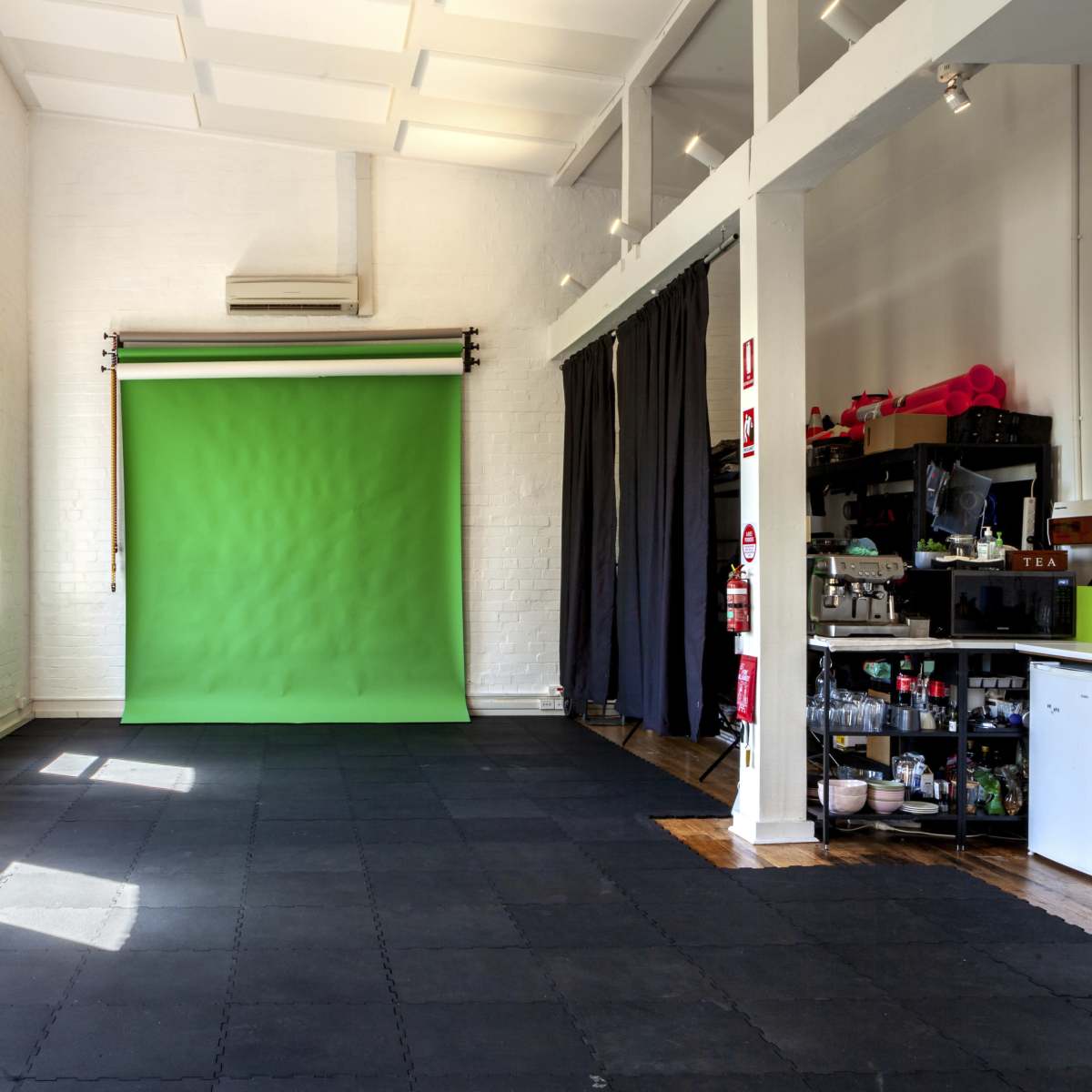 A spacious studio with a green backdrop, large windows allowing natural light, and a kitchenette area on one side.
