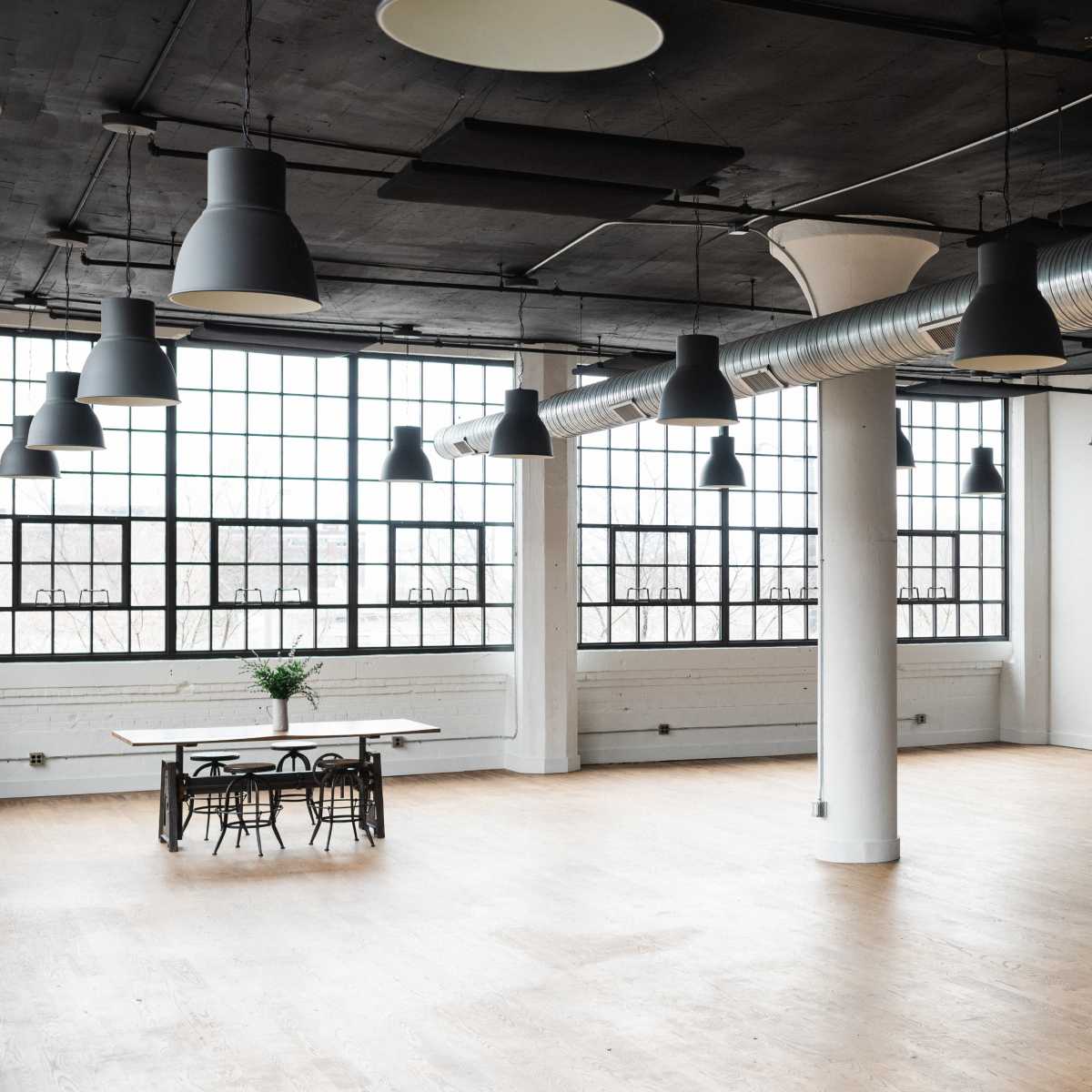 A spacious, modern interior with large windows, exposed ductwork, and a wooden table surrounded by chairs.