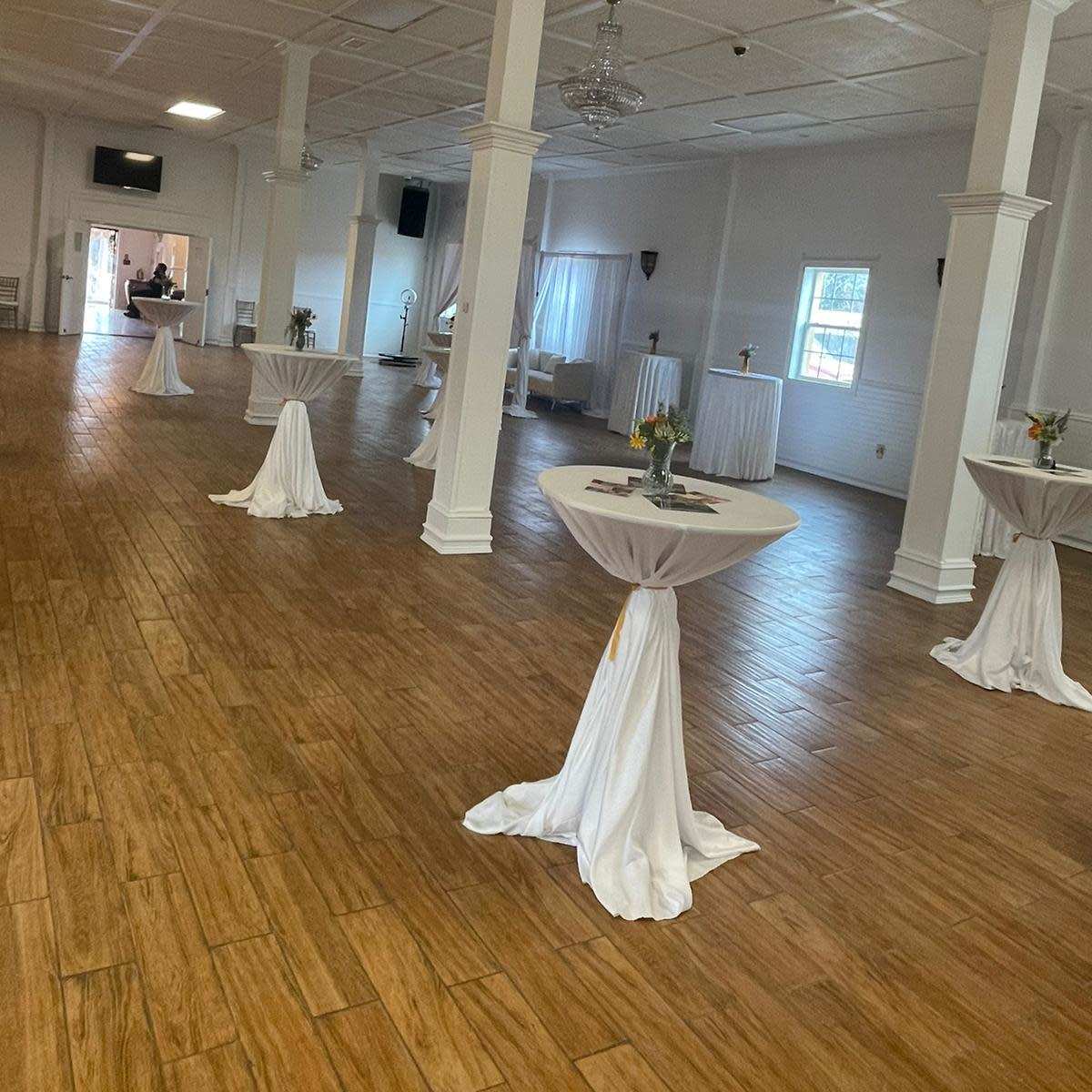 An indoor event space with polished wooden floors, round tables dressed in white tablecloths, and a few floral centerpieces arranged on the tables.