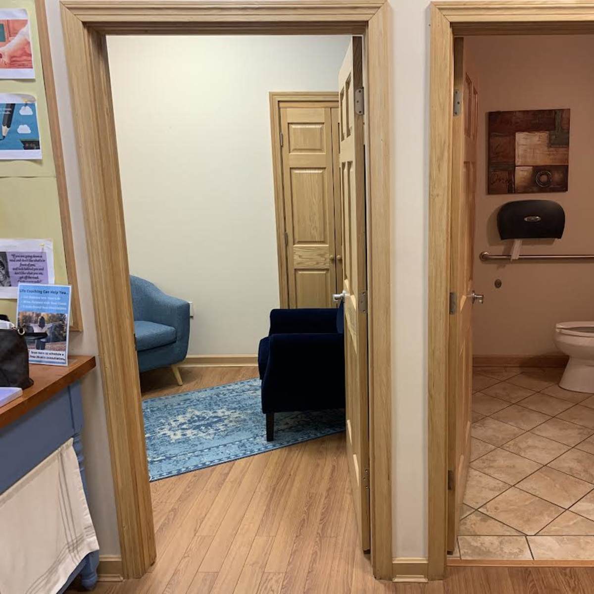 A hallway with two doorways, one leading to a small sitting area with a blue chair and the other leading to a bathroom with a toilet and sink.