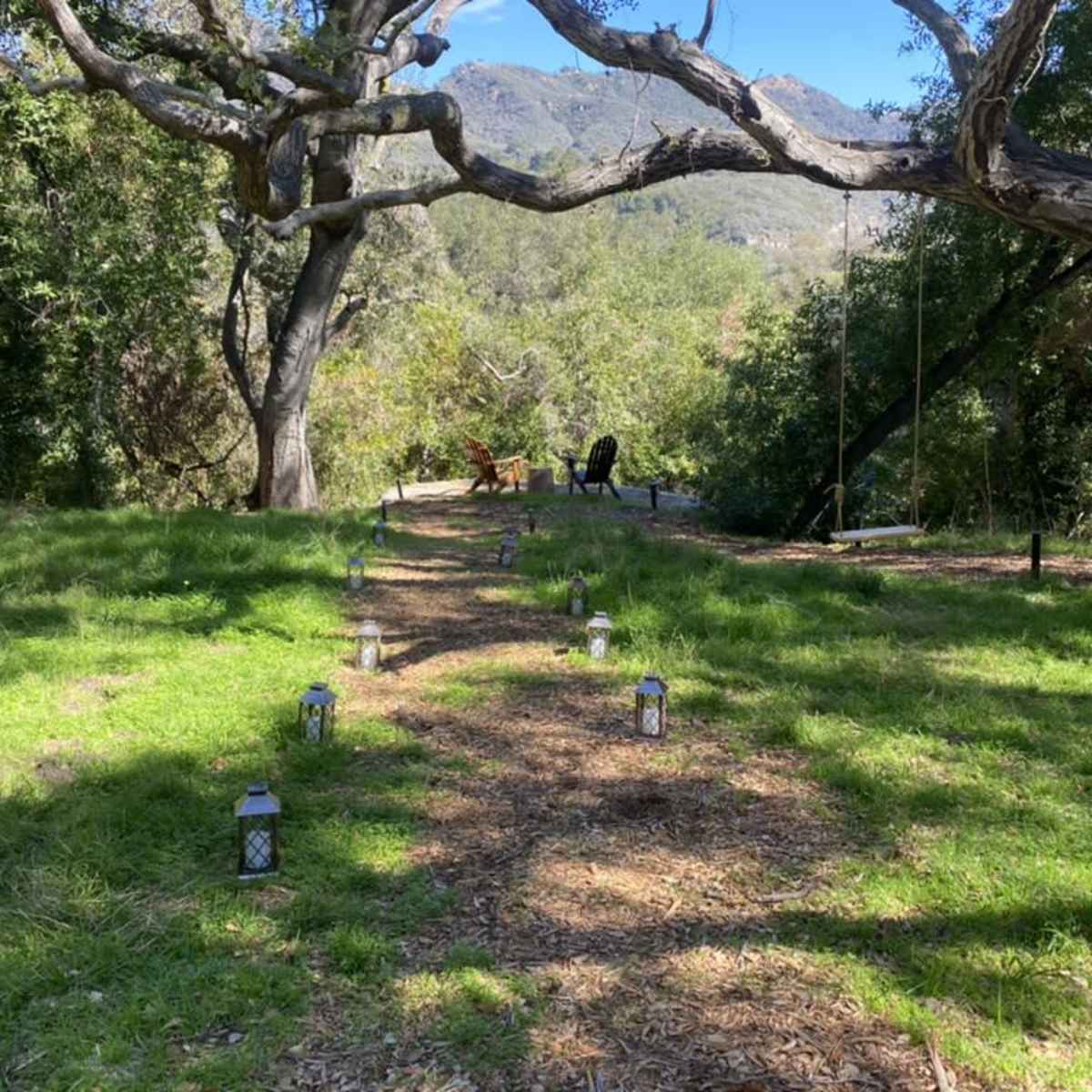 A pathway lined with lanterns leads to a seating area beneath large trees and mountains in the background.