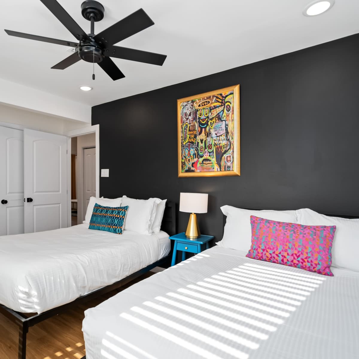 A bedroom with two single beds, a vibrant piece of artwork on the wall, and a ceiling fan, with an open door leading to another room.