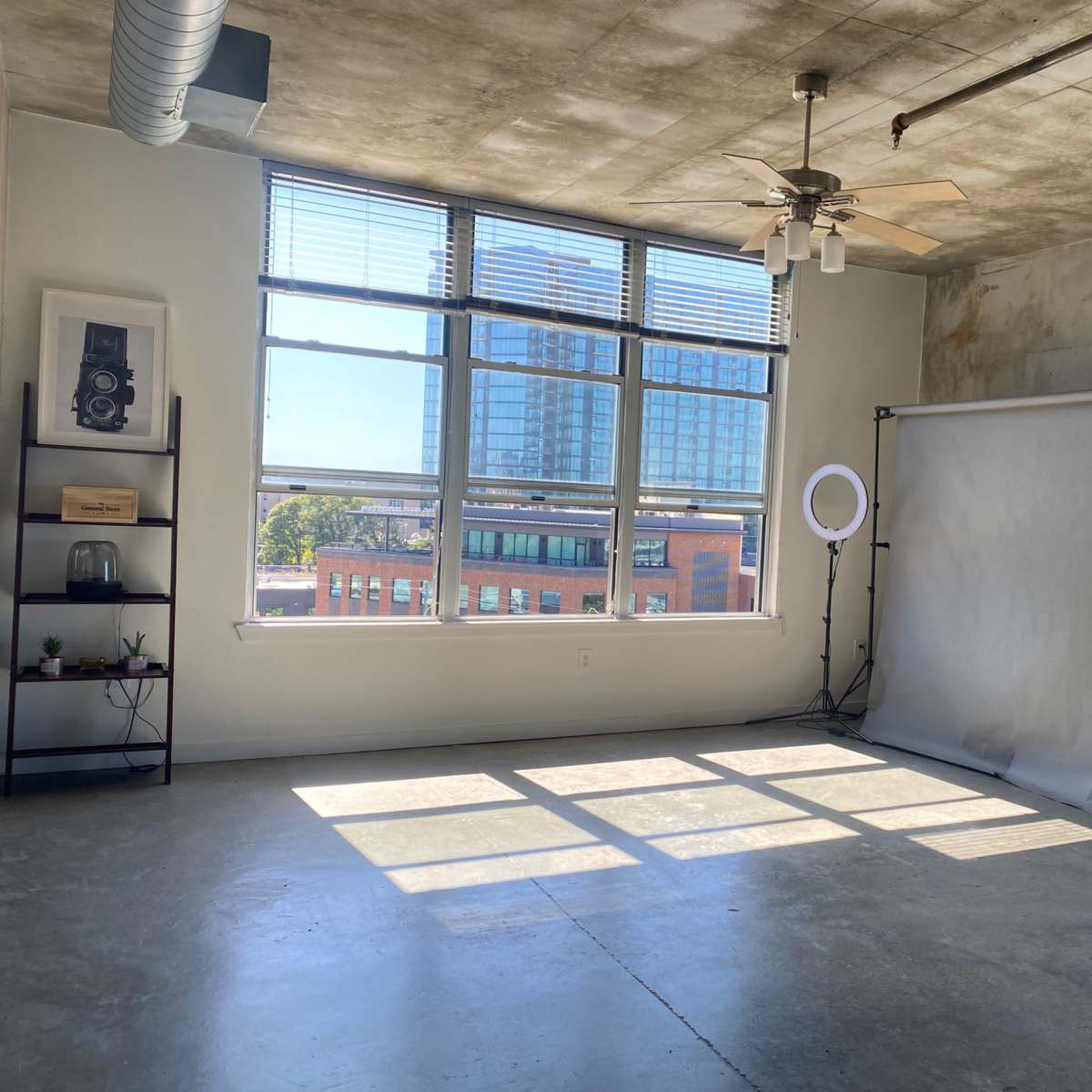 A spacious, well-lit room features large windows, a ceiling fan, and a backdrop setup against a concrete wall.