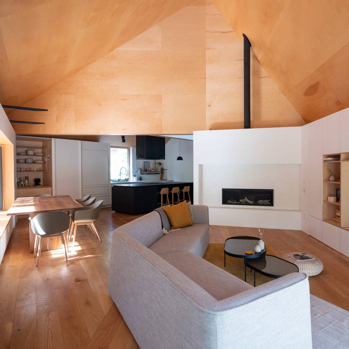 A modern, open-concept living room with a large sofa, a dining area, and a sleek kitchen, all featuring wooden accents and a minimalist design.