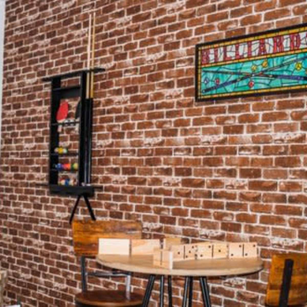 A cozy room features a brick wall with a stained glass window, a round table with wooden chairs, and various game pieces displayed on a shelf.