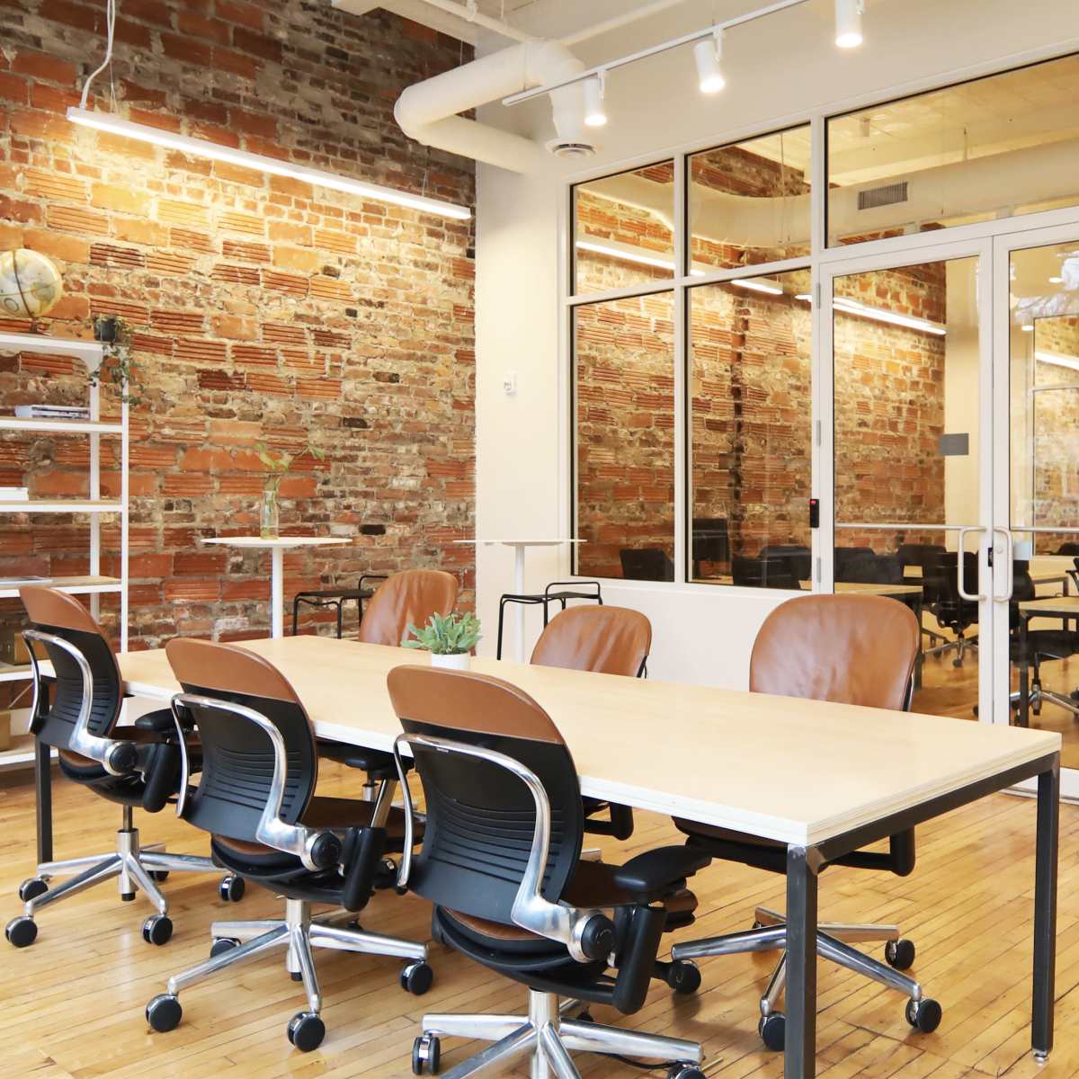 A modern office space with a long table and several ergonomic chairs, featuring exposed brick walls and large windows.