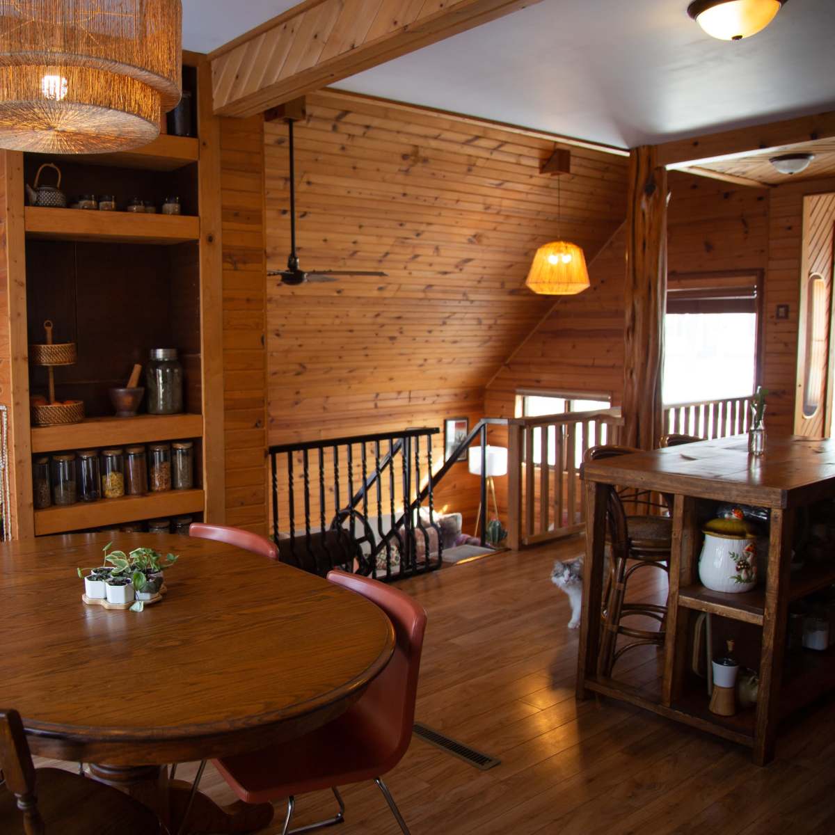 A wooden interior of a home featuring a dining table with chairs, a bookshelf, and an open staircase.