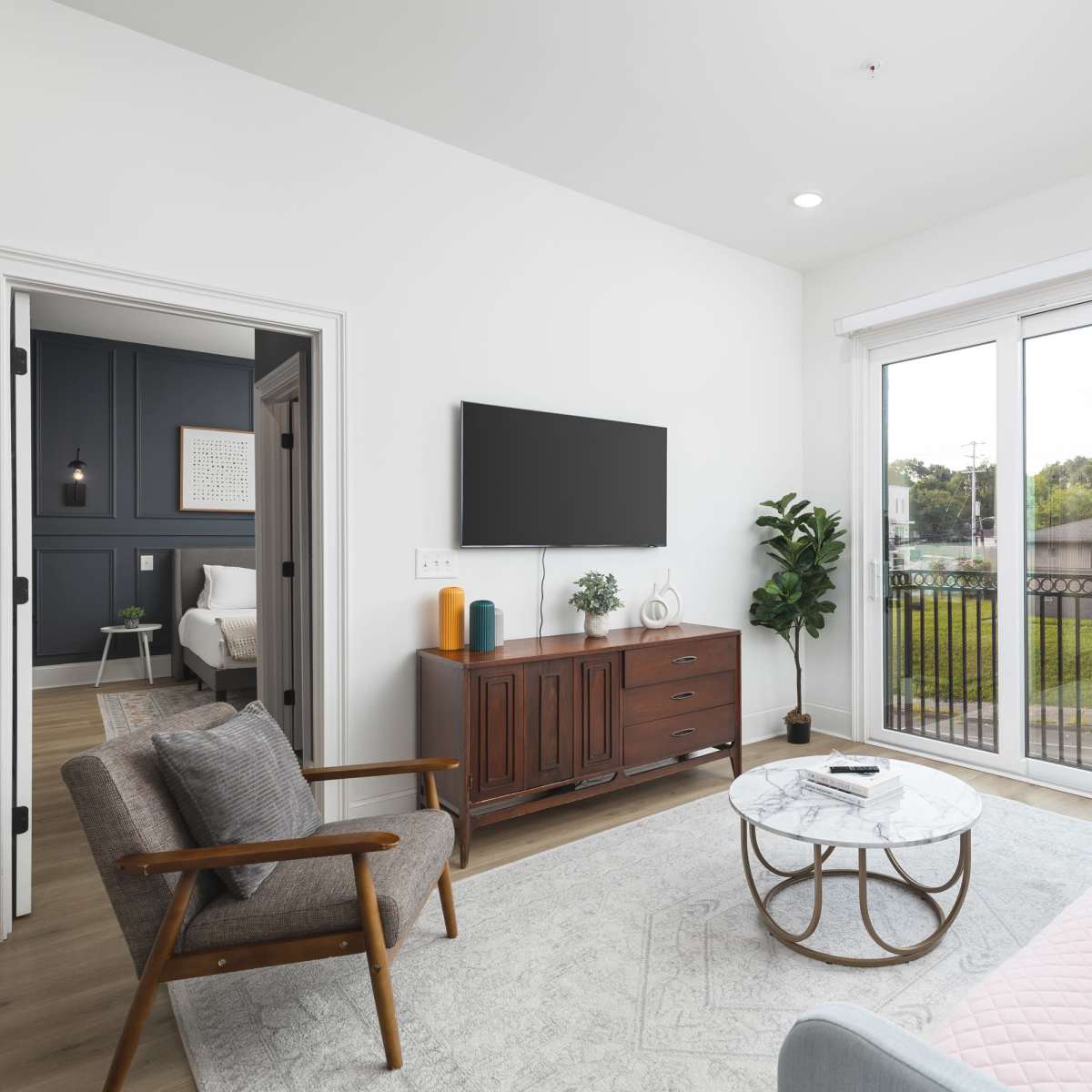 A modern living room features a light-colored sofa, a glass coffee table, and large sliding doors that open to a balcony, with a view of the outdoors visible through the glass.