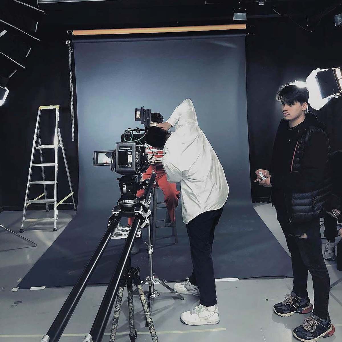 A person in a white jacket operates a camera on a tracking dolly while another individual stands nearby in a dimly lit studio with a backdrop and lighting equipment.