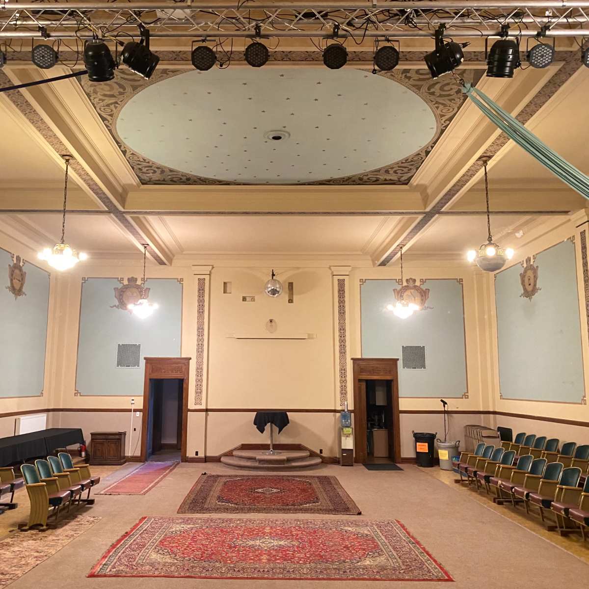A spacious hall with rows of green chairs, a large decorative ceiling, and a stage at the front, flanked by two open doors.