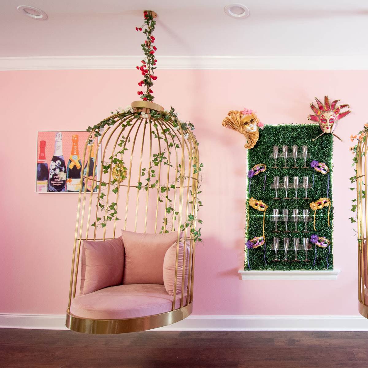 Two hanging birdcage chairs adorned with flowers and greenery, flanking a wall decorated with a vertical display of glass champagne flutes and colorful masks against a pink backdrop.