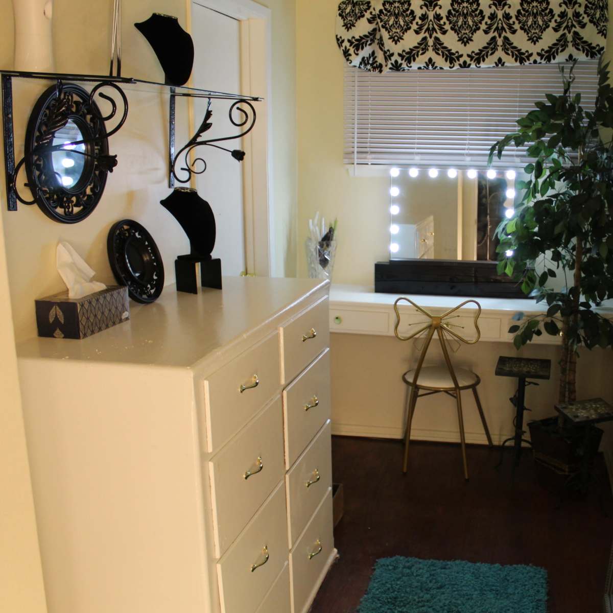 A small dressing area with a mirrored vanity table, a chair, a decorative plant, and a dresser with multiple drawers.
