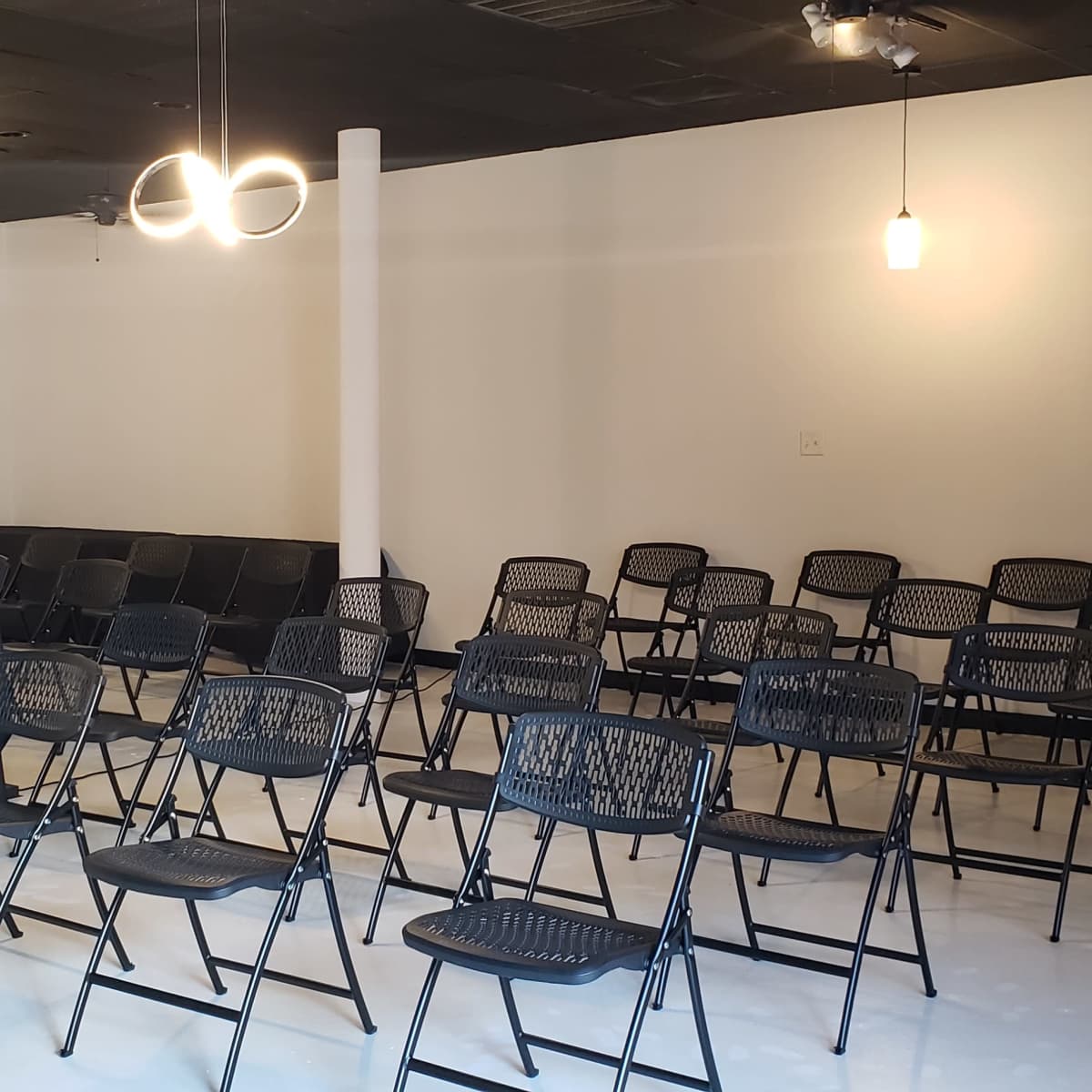 A spacious room with black folding chairs arranged in rows and a tall table in the center, illuminated by modern pendant lights.