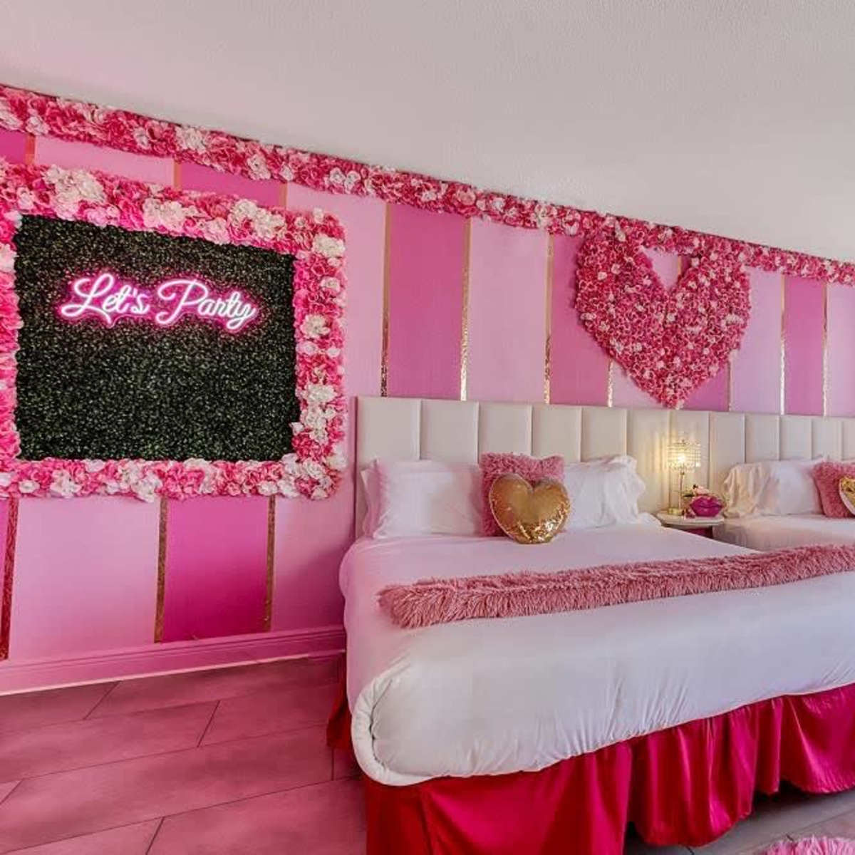 A brightly decorated hotel room with two beds, pink walls, floral accents, and a neon sign reading "Let's Party."