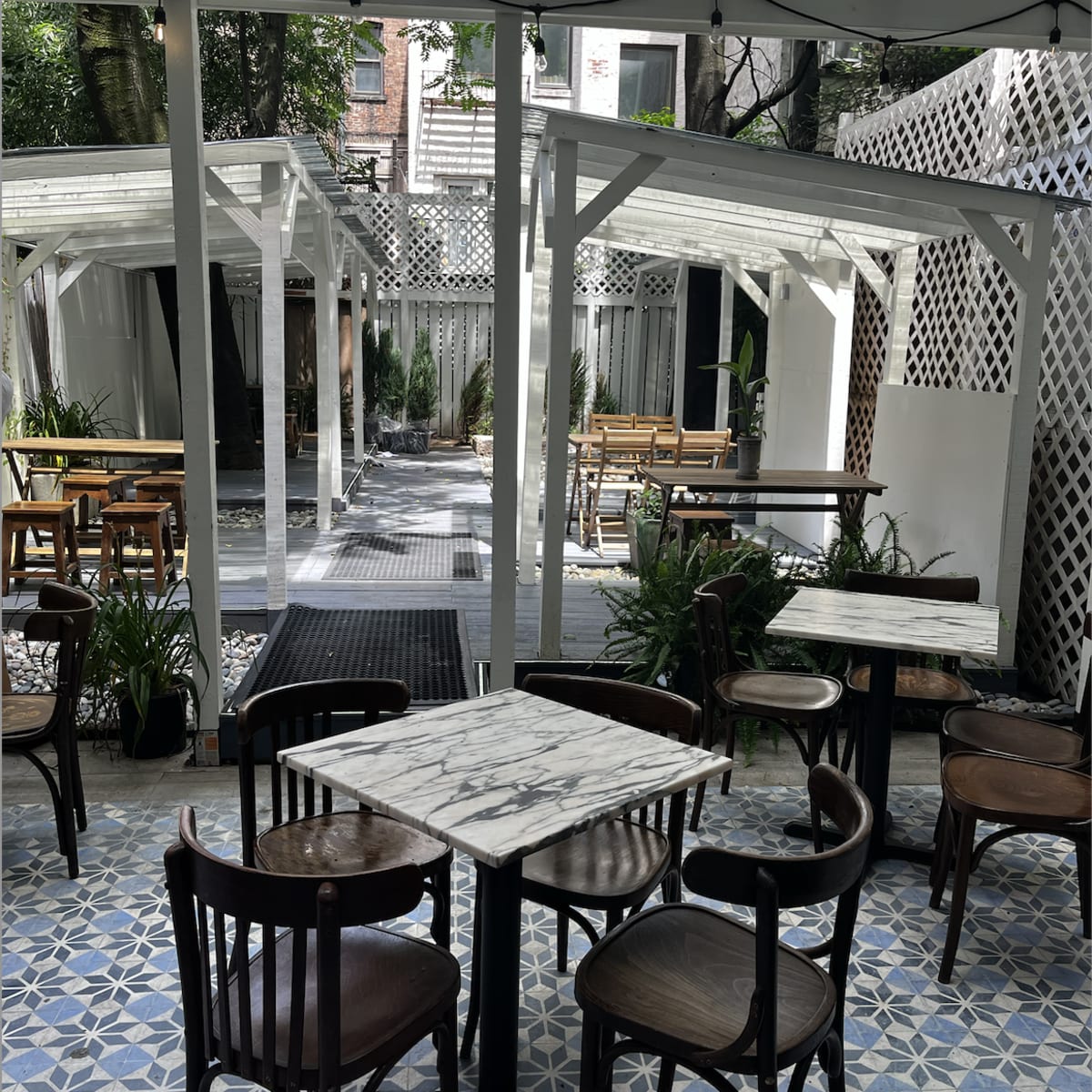 A patio area of a café with wooden tables and chairs, surrounded by greenery and an open, lattice-covered structure.