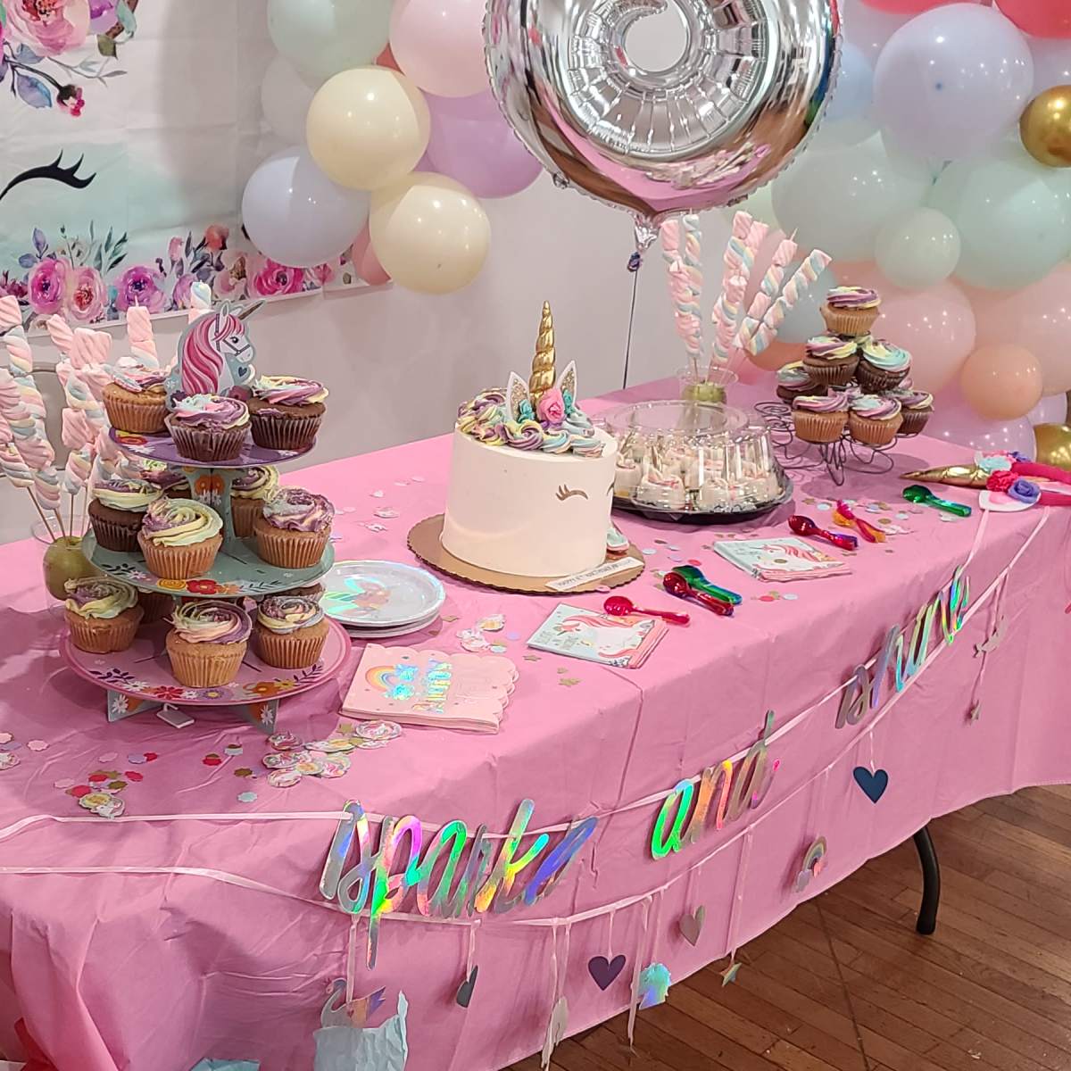 A decorated party table featuring a unicorn-themed cake, colorful cupcakes, and various candy items surrounded by balloons.