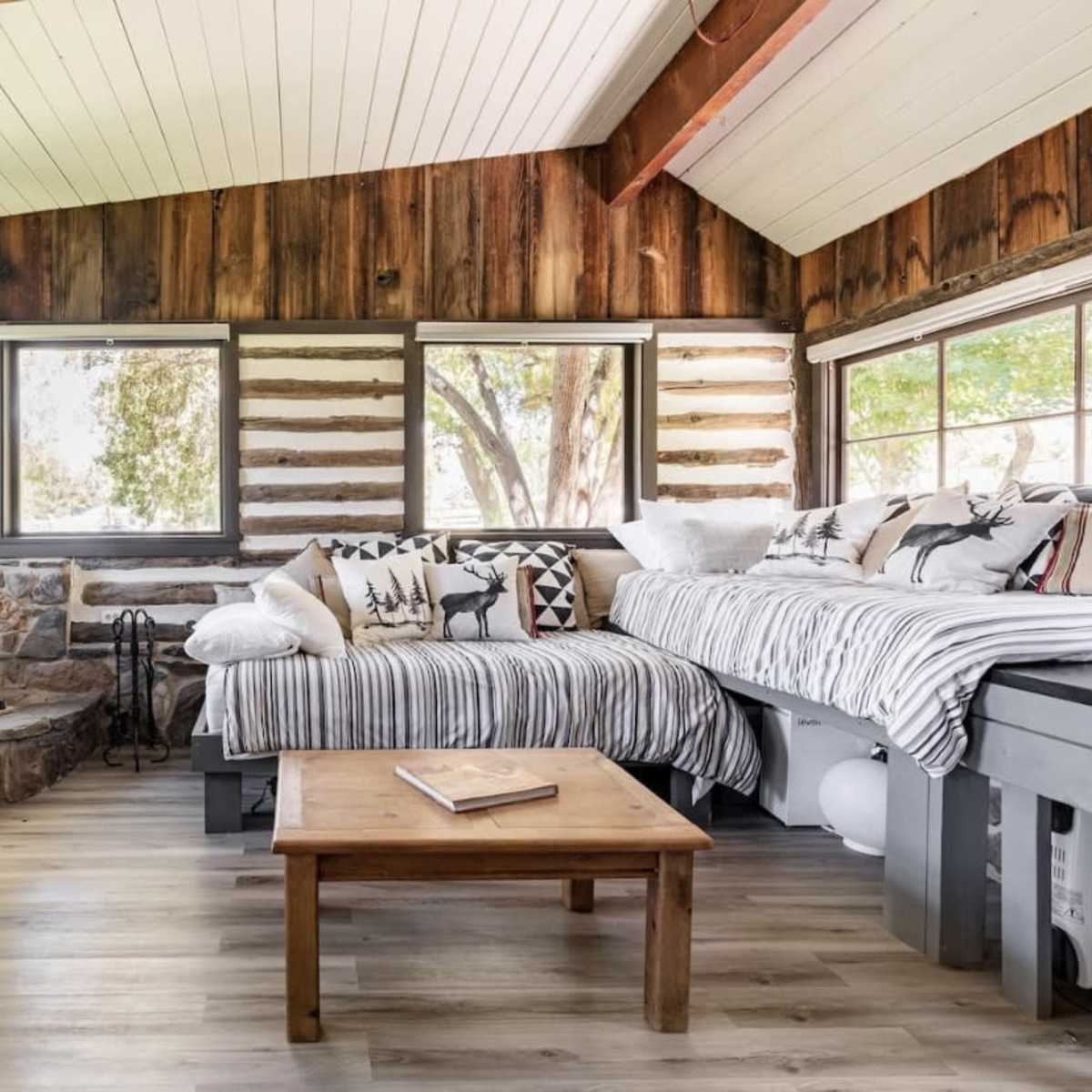 A cozy cabin interior featuring a wood stove, wooden walls, a coffee table, and a seating area with patterned pillows.