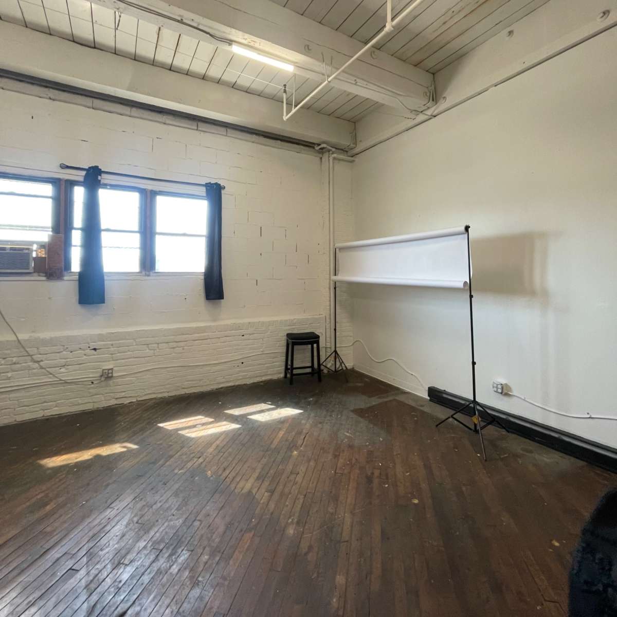 A sparse room with wooden floors, white walls, two windows dressed with black curtains, and a folding screen positioned against one wall.