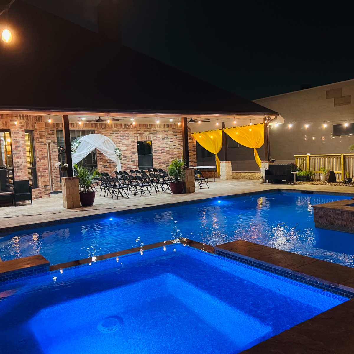 A well-lit outdoor pool area with a hot tub, surrounded by lounge chairs and decorated with string lights and yellow drapes.