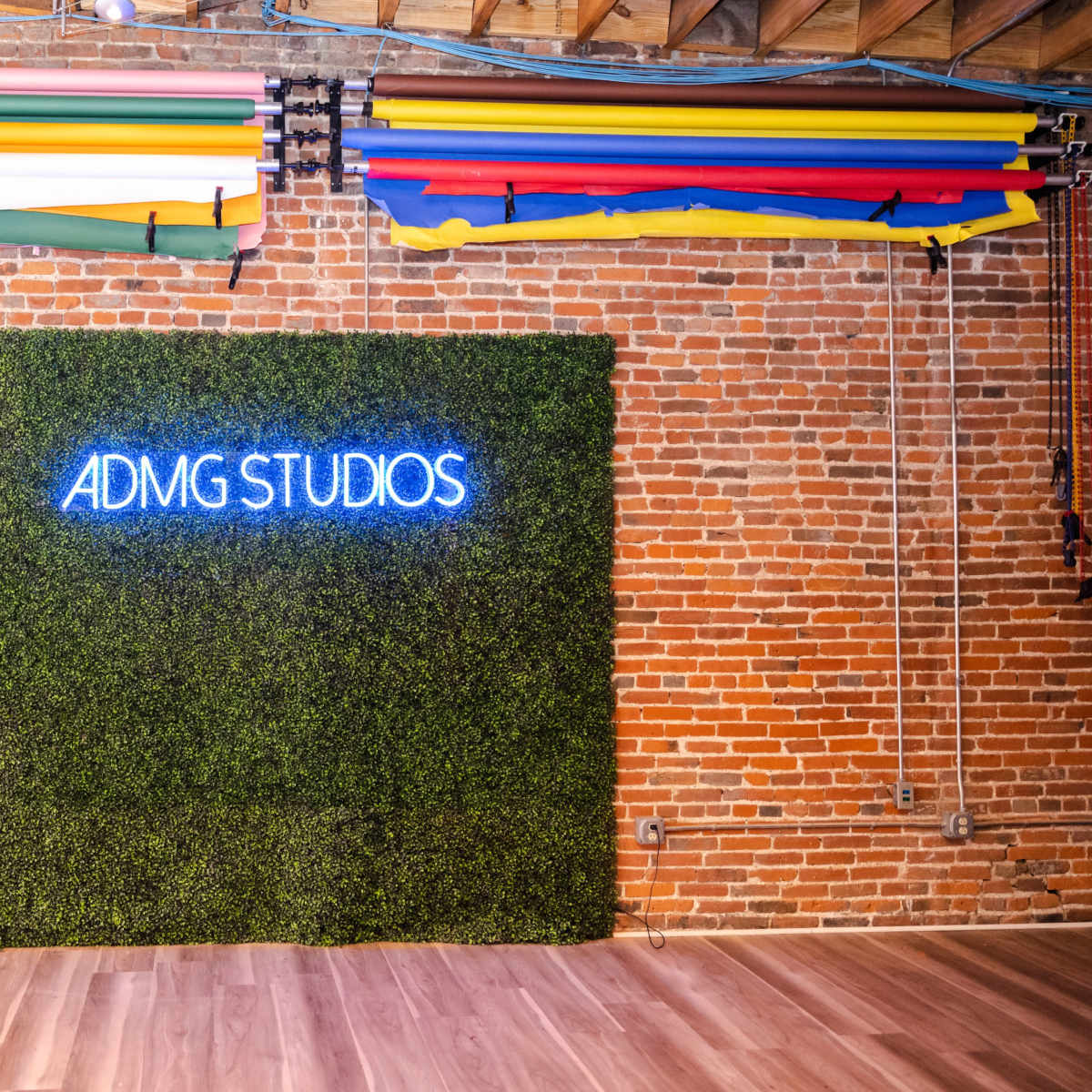 A brick wall with a green moss-like backdrop and the neon sign "ADMG STUDIOS" illuminated above it, complemented by colorful rolls of backdrops hanging overhead.