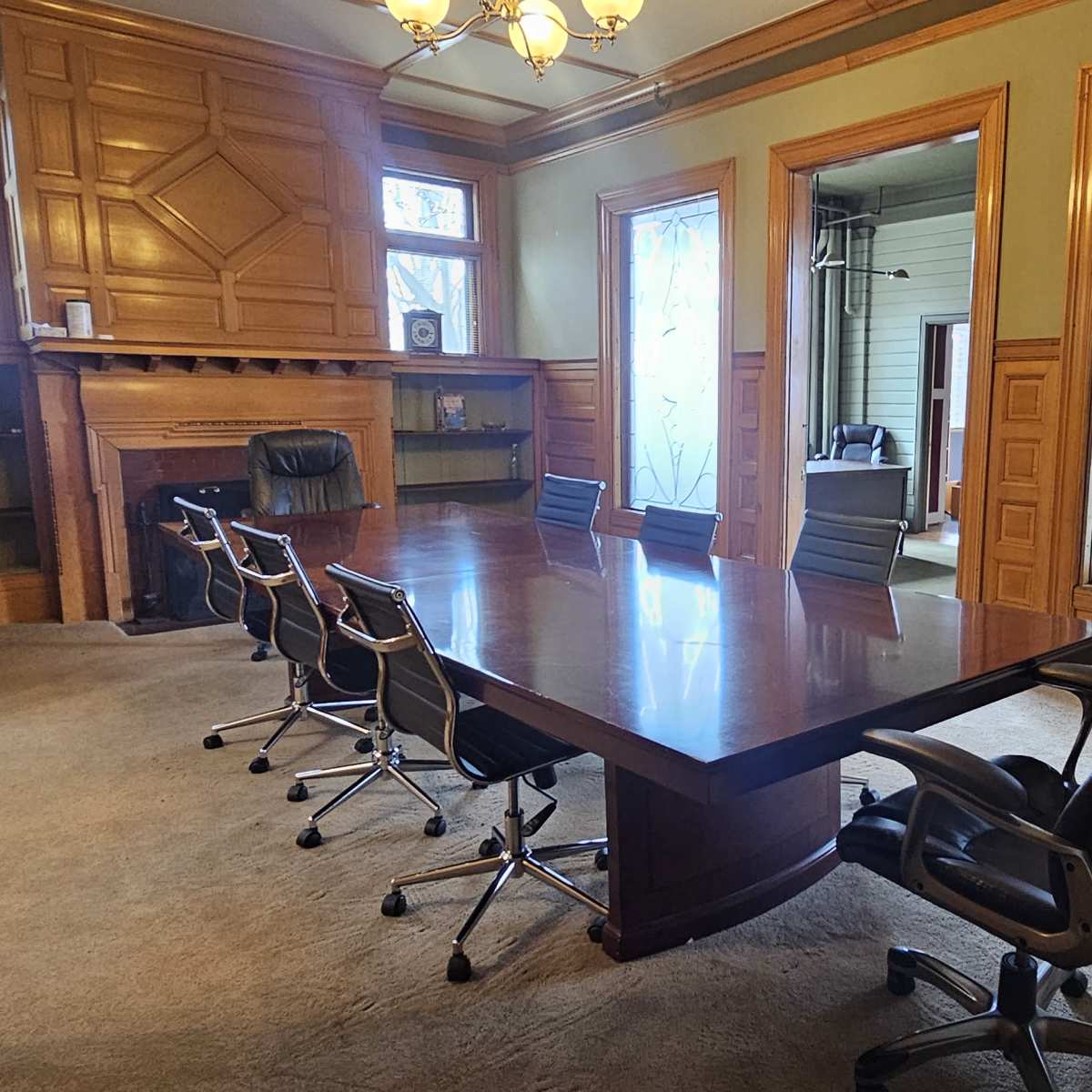 A wooden conference room with a large table surrounded by six chairs, a fireplace, and windows featuring decorative glass.