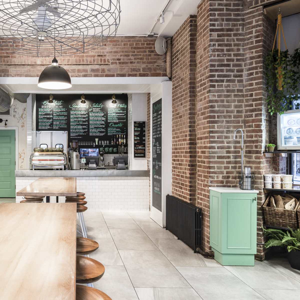 A modern café interior features a long wooden table, black chairs, exposed brick walls, and a counter with a coffee machine and menu overhead.
