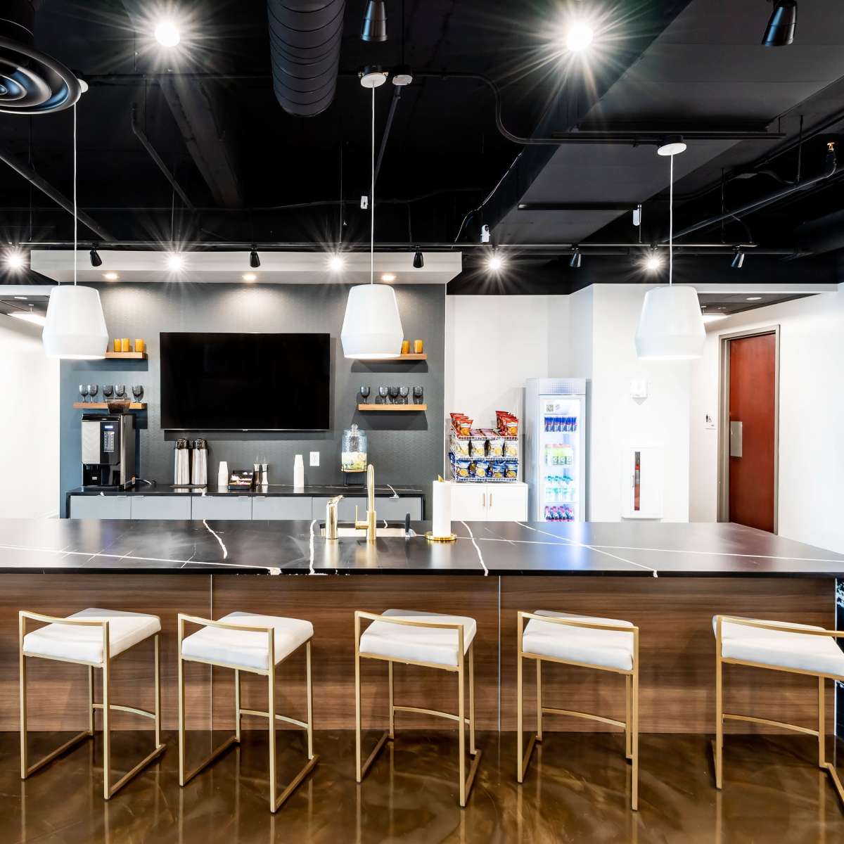 A modern kitchen area with a long, dark countertop and seven white barstools under it, featuring a television and vending machine in the background.