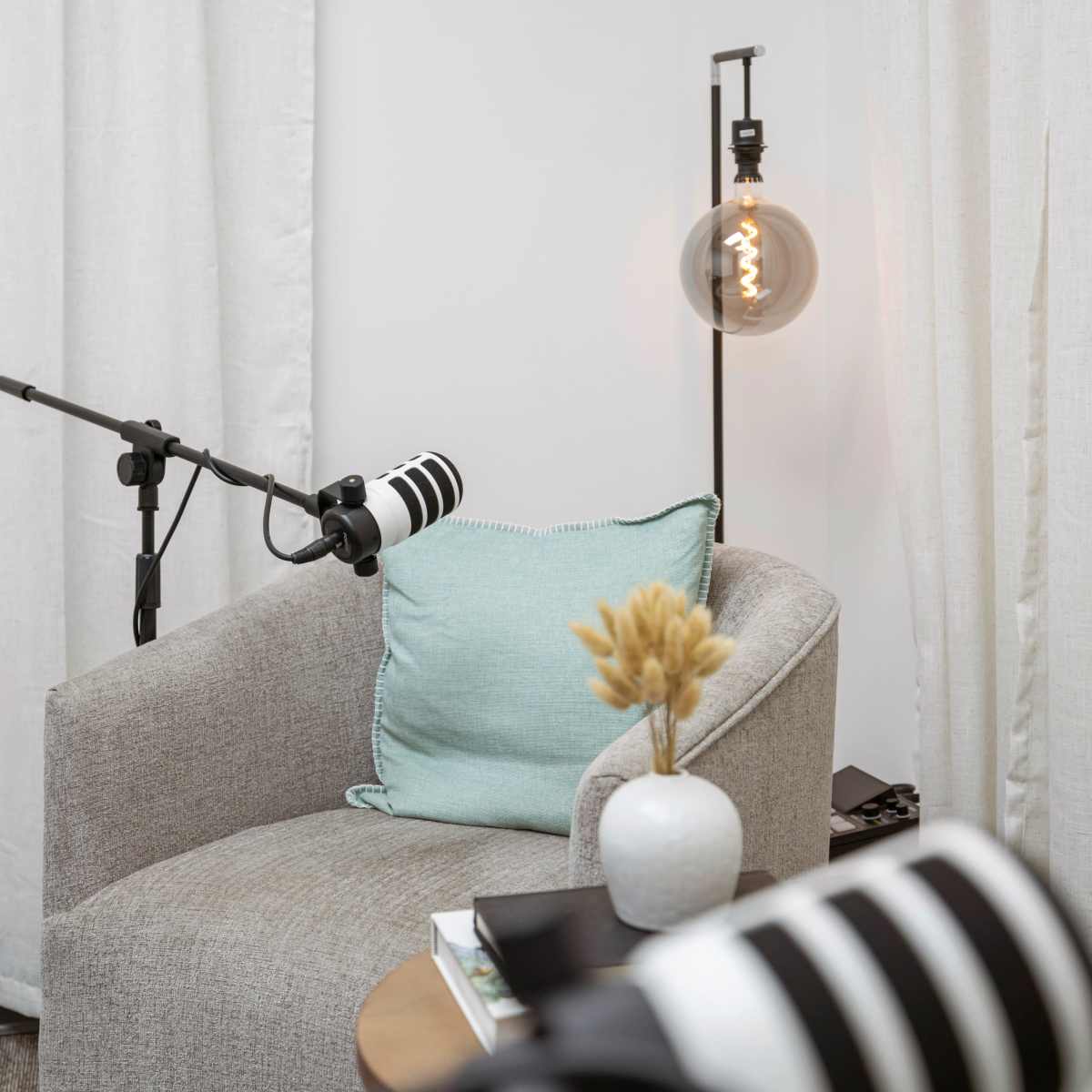 A microphone setup is positioned next to a cozy chair adorned with a light blue pillow, accompanied by a small vase of dried flowers and a lamp with an exposed bulb.