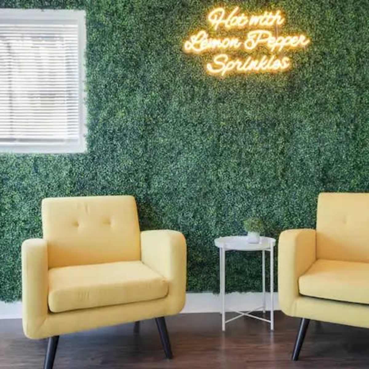 Two yellow armchairs are positioned facing each other in front of a green wall adorned with a neon sign that reads "Hot with Lemon Pepper Sprinkles."