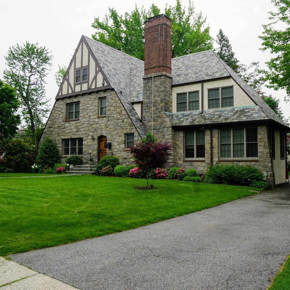 A large, stone Tudor-style house with a slate roof, situated on a manicured lawn alongside a paved driveway.