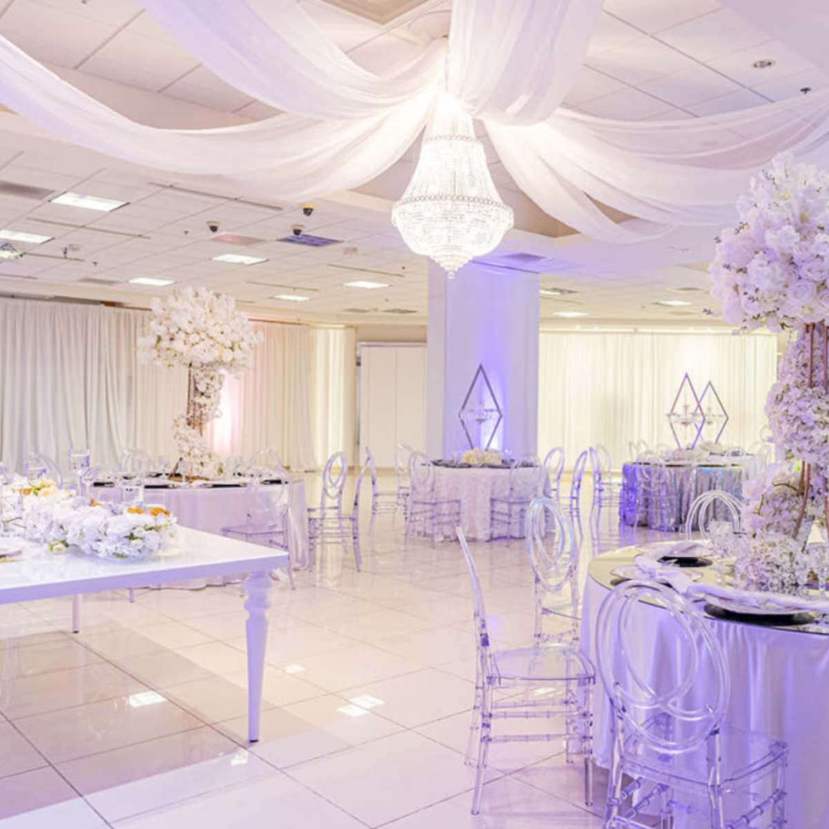 An elegantly decorated banquet hall with white table settings, floral centerpieces, and draped fabric overhead.