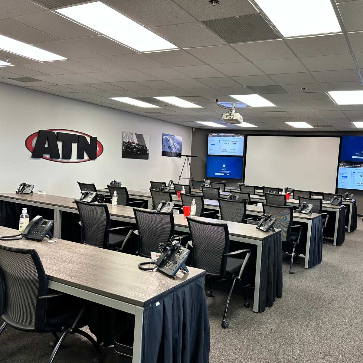 A training room features rows of desks with phones, large screens displaying information, and a projector, all set up for a corporate seminar.