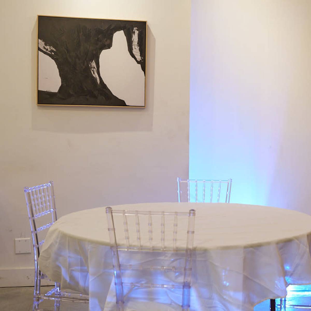 A round table with a white tablecloth is set in a room with clear acrylic chairs, accompanied by an artwork on the wall.