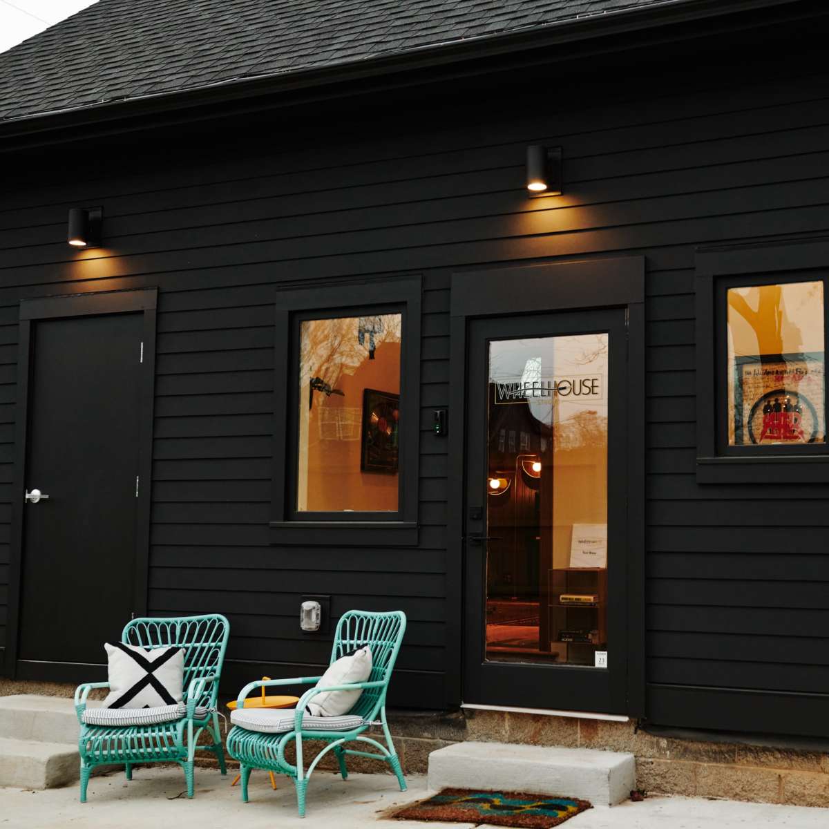 A black wooden house exterior featuring a main entrance door and two windows, with green wicker chairs placed outside on a concrete walkway.