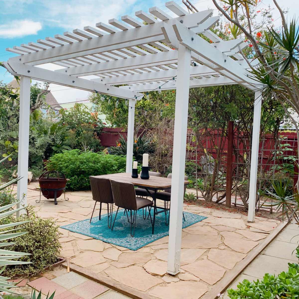 A patio area with a white pergola, a wooden table set with chairs, and surrounded by lush greenery and stone pathways.