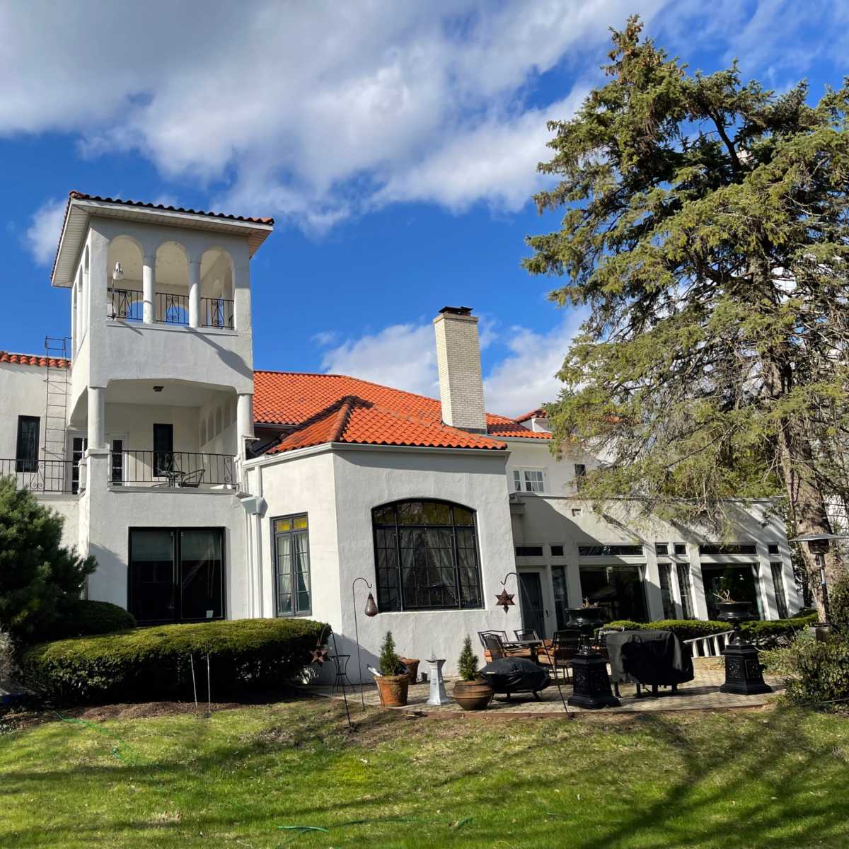 A large, two-story white house with a red tile roof and a tower overlooks a manicured lawn and garden.