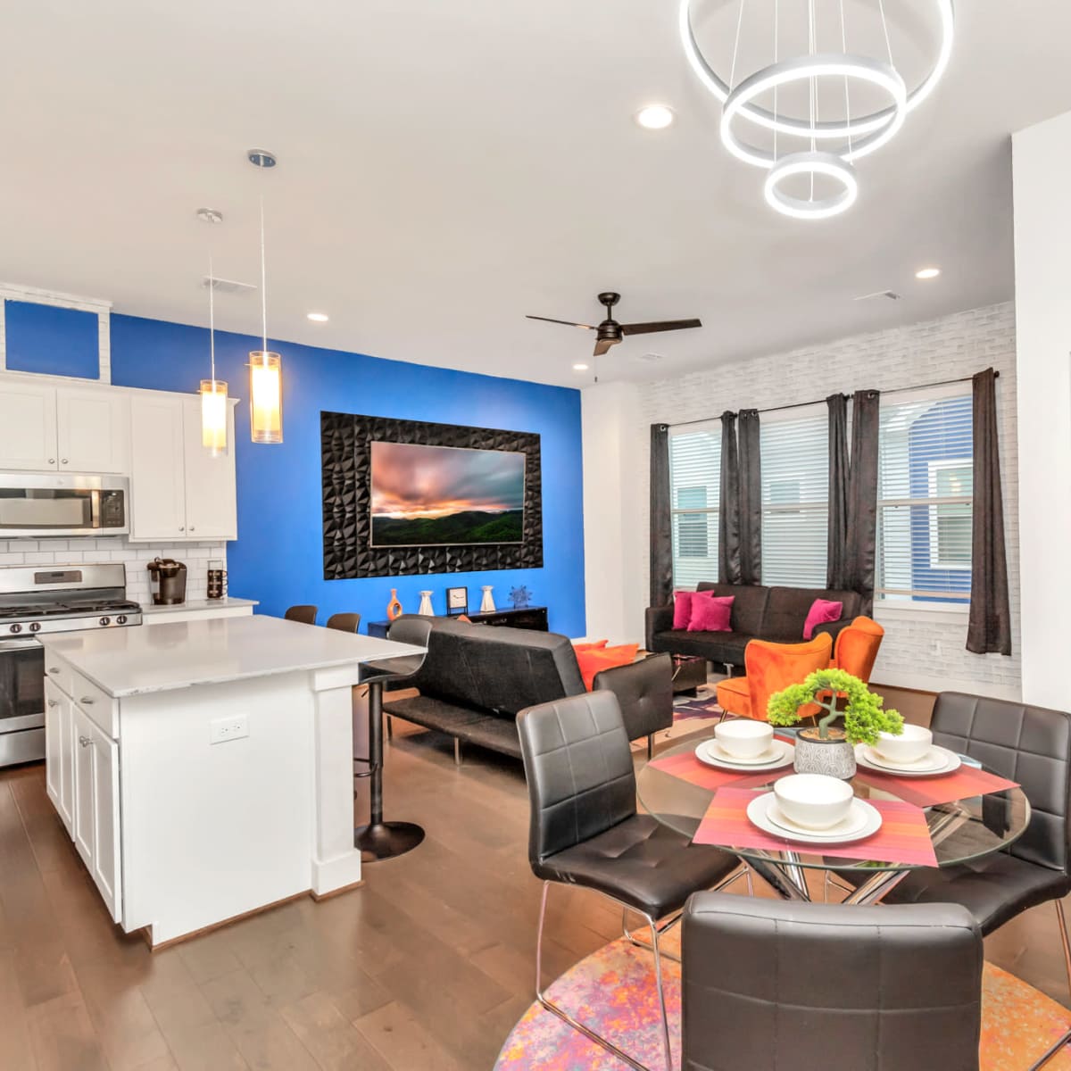 A modern open-concept living space featuring a kitchen area with white cabinetry, a dining table set for four, and a living room with colorful seating and a large television on the wall.