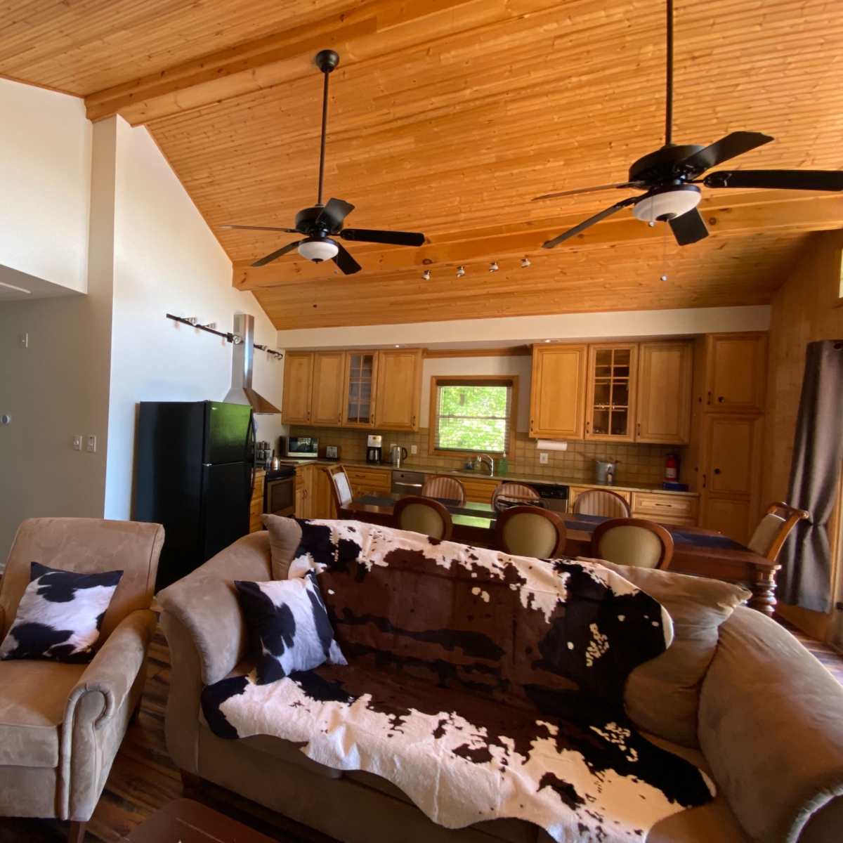 A spacious living room with a high wooden ceiling, featuring a sofa draped with a cowhide blanket, a kitchen area in the background, and a dining table.