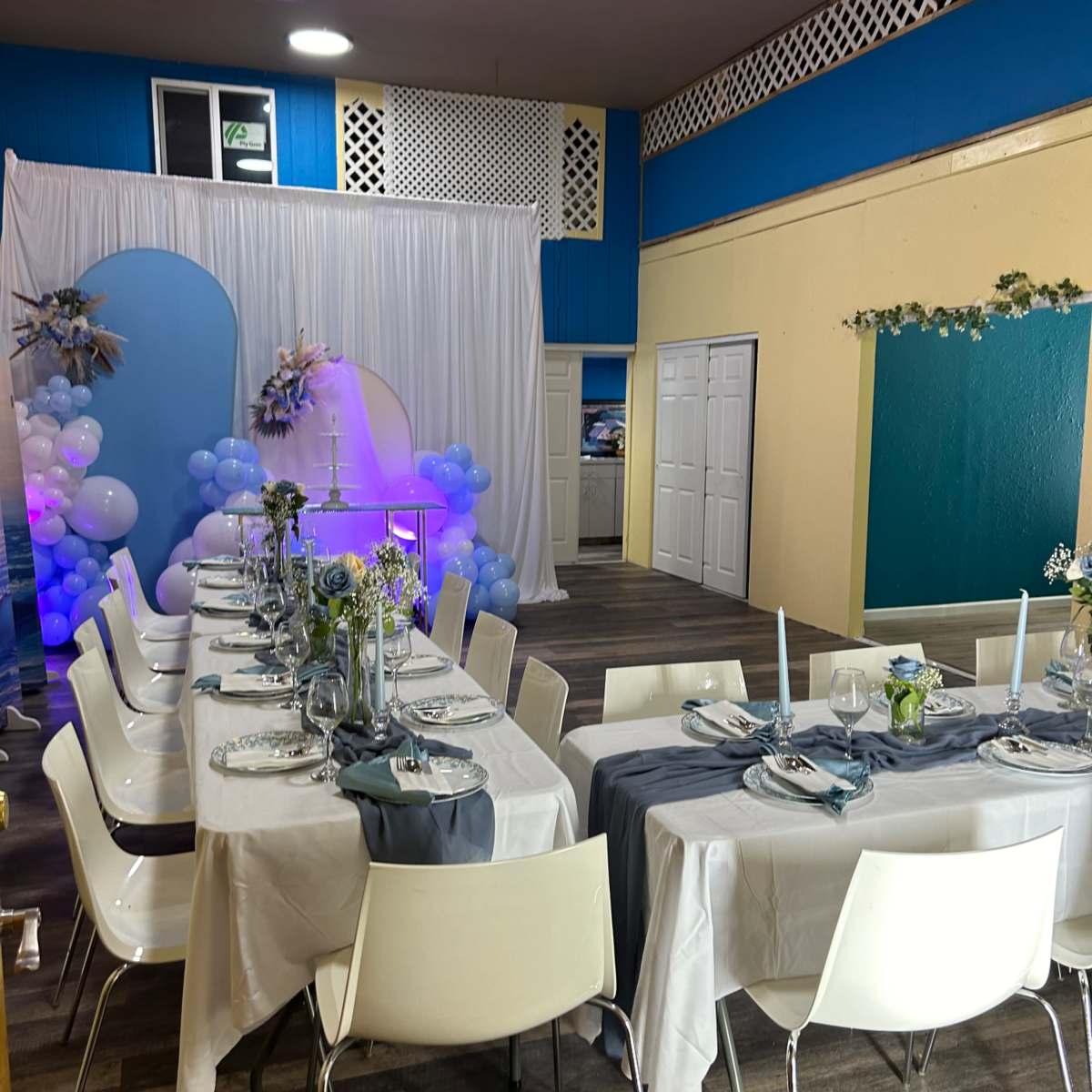 A decorated event space with two tables set for dining, featuring blue and white balloons, floral arrangements, and table settings with plates and glasses.